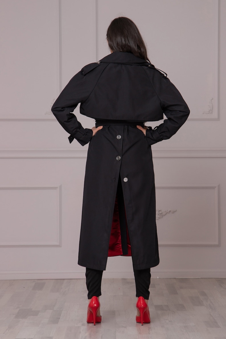 Puede incluir: Una gabardina negra con cintur&oacute;n y botones. La gabardina tiene un forro rojo y se muestra desde atr&aacute;s.
