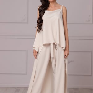May include: A beige sleeveless top with a draped front and a matching pair of wide-leg pants with a tie at the waist. The pants have a long, flowing skirt overlay.