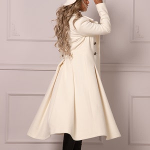 May include: A white long coat with a flared skirt and a white hat. The coat has a double breasted closure and a high collar. The model is wearing black boots.