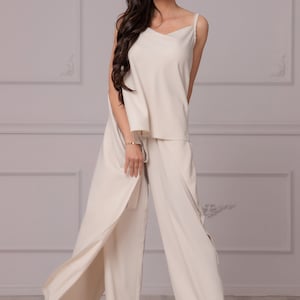 May include: A woman wearing a cream-colored pantsuit with a wide-leg, flowing pant style. The pantsuit has a sleeveless top with a V-neckline. The woman is wearing red heels.