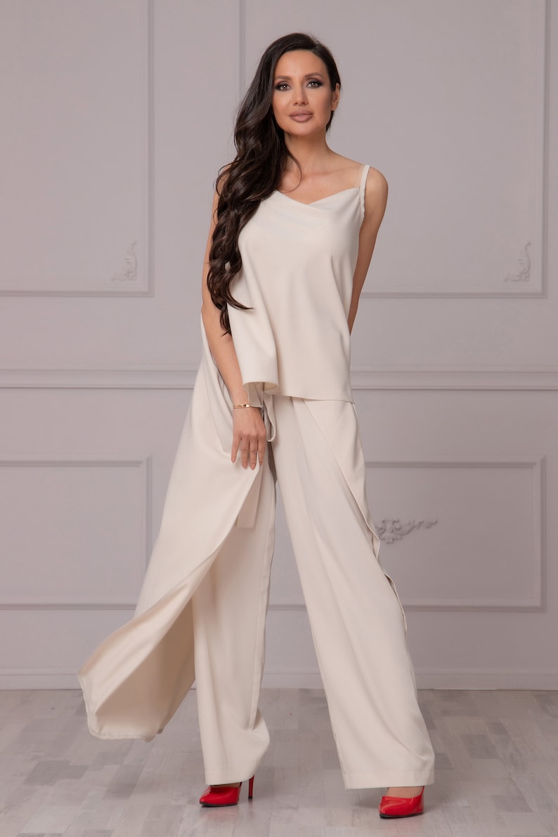 May include: A woman wearing a cream-colored pantsuit with a wide-leg, flowing pant style. The pantsuit has a sleeveless top with a V-neckline. The woman is wearing red heels.