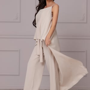 May include: A woman wearing a beige pantsuit with a long, flowing skirt. The pantsuit has a wide-leg silhouette and a tie at the waist. The woman is wearing red high heels.