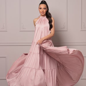K&ouml;nnte beinhalten: Eine Frau tr&auml;gt ein langes, flie&szlig;endes, rosafarbenes Maxikleid mit mehreren Lagen und einem Halterneck. Das Kleid ist aus einem weichen, seidenartigen Stoff gefertigt und hat eine flie&szlig;ende, lockere Passform.