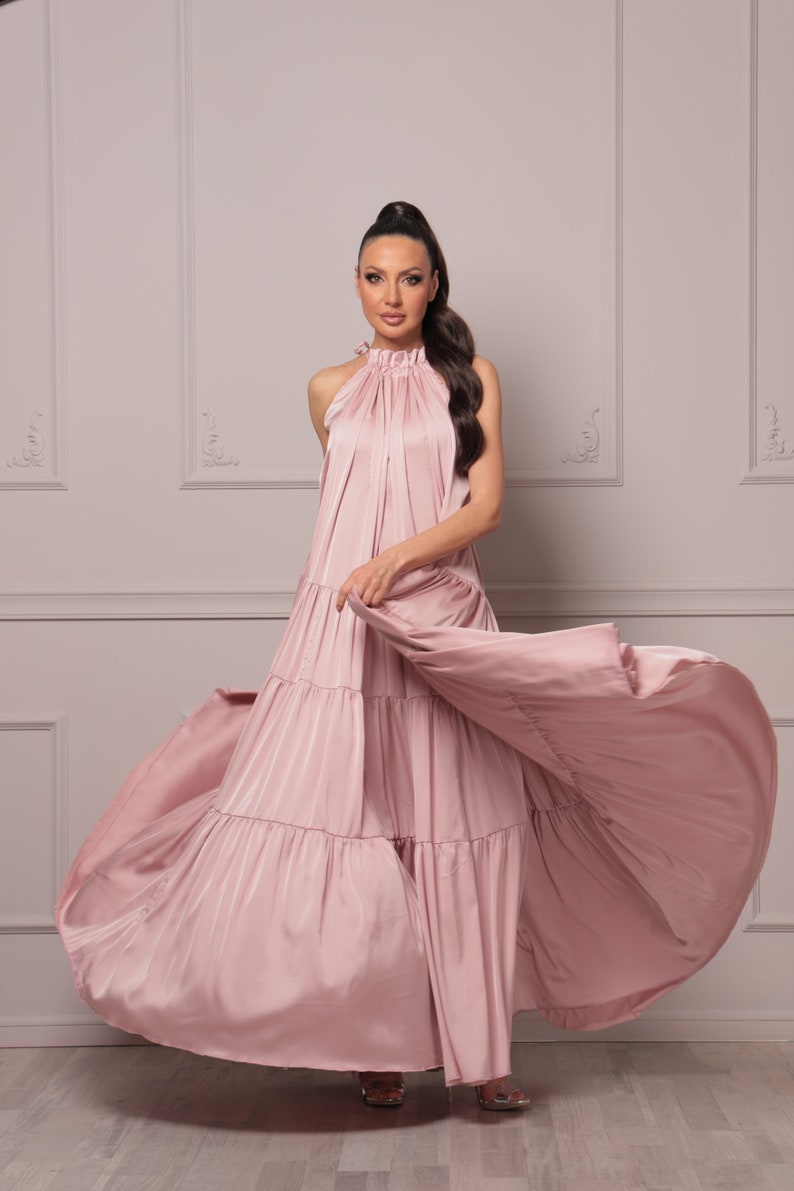 K&ouml;nnte beinhalten: Eine Frau tr&auml;gt ein langes, flie&szlig;endes, rosafarbenes Maxikleid mit mehreren Lagen und einem Halterneck. Das Kleid ist aus einem weichen, seidenartigen Stoff gefertigt und hat eine flie&szlig;ende, lockere Passform.