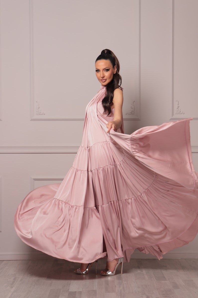 K&ouml;nnte beinhalten: Eine Frau in einem langen, flie&szlig;enden rosa Kleid mit mehreren Lagen. Das Kleid ist aus einem weichen, seidenartigen Stoff und hat einen hohen Ausschnitt. Die Frau steht auf einem hellen Holzboden und ihre Haare sind zu einem Pferdeschwanz gebunden.