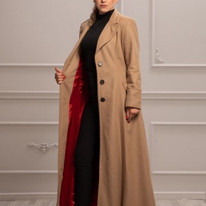 K&ouml;nnte beinhalten: Ein langer, beiger Mantel mit rotem Futter. Der Mantel hat einen Kragen, einen Knopfverschluss und lange &Auml;rmel. Das Model tr&auml;gt eine schwarze Hose und rote Schuhe mit Absatz.