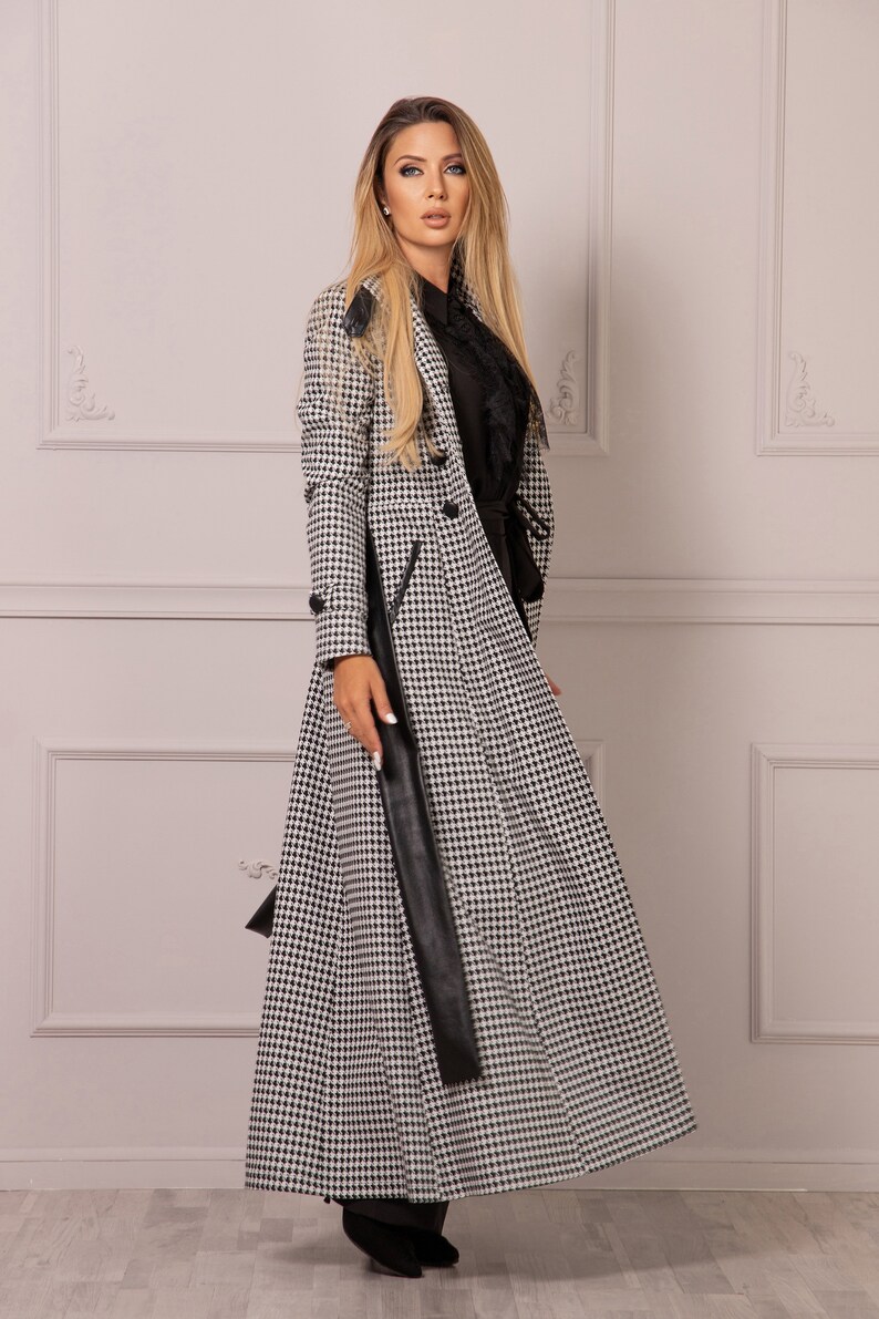 May include: A long black and white houndstooth coat with a black leather belt and a black collar. The coat has a double breasted closure and a long, flowing skirt.