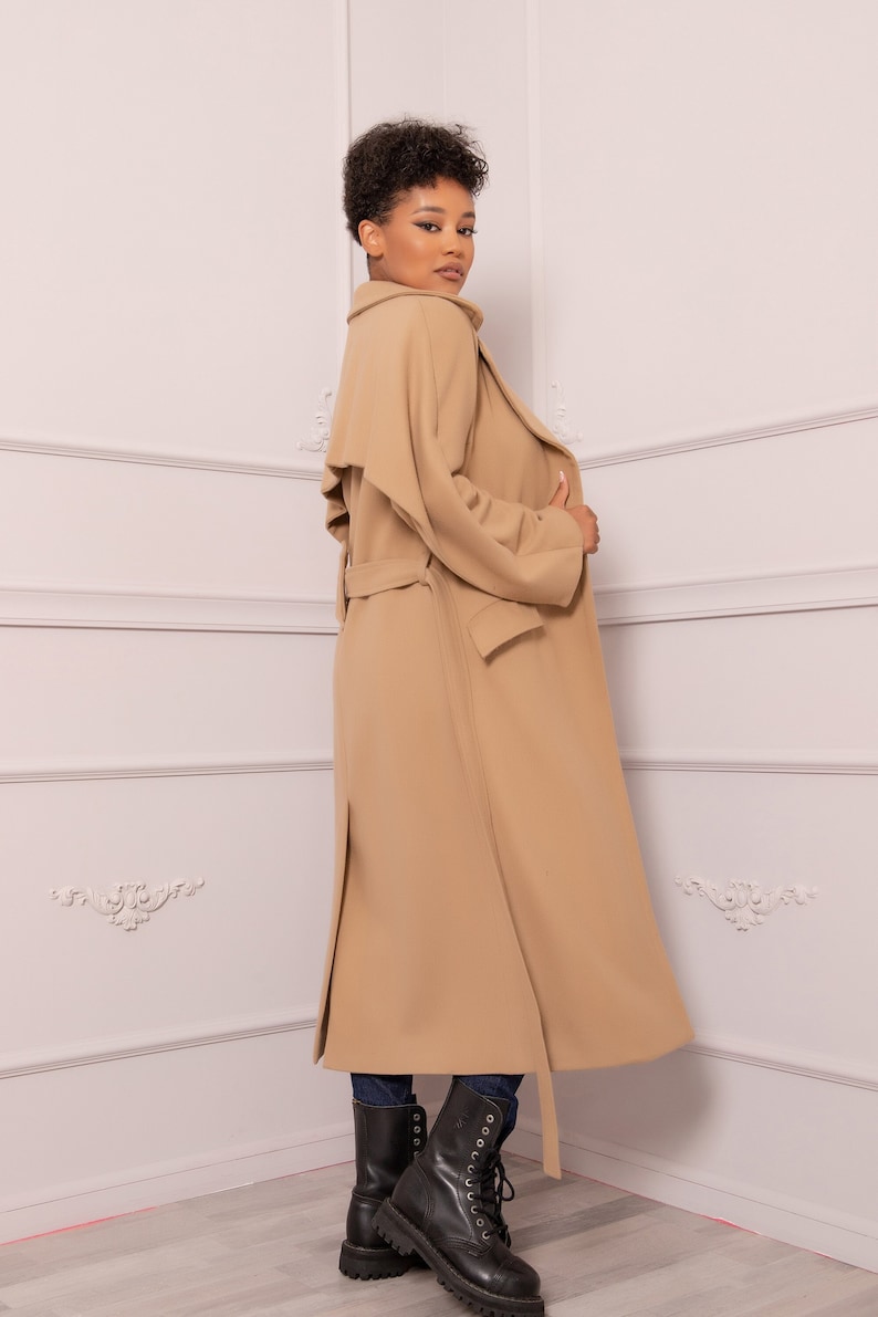 Op de afbeelding: Een lange, beige trenchcoat met een riem en een kraag die omhoog of omlaag gedragen kan worden. De jas is gemaakt van een zachte, comfortabele stof en heeft een ontspannen pasvorm.