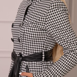 May include: A black and white houndstooth patterned coat with a black leather collar, belt, and trim. The coat has a double breasted closure with black buttons and a long sleeve.