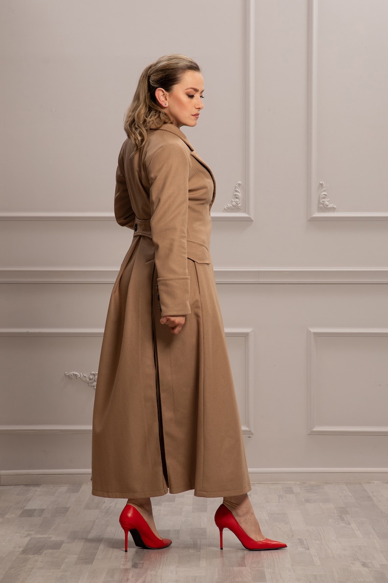 K&ouml;nnte beinhalten: Ein langer, brauner Mantel mit hohem Kragen und einreihiger Knopfleiste. Der Mantel hat einen taillierten Oberk&ouml;rper und einen flie&szlig;enden Rock mit einem Schlitz im R&uuml;cken. Das Model tr&auml;gt rote High Heels.