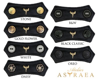 May include: Six black fabric face mask extenders with different colored buttons. The extenders feature a gold embroidered butterfly design. The buttons include gold, black, white, and clear rhinestones. The text "STONE", "GOLD FLOWER", "B&W", "BLACK CLASSIC", "WHITE", "DAISY", and "OREO" are printed below each extender. The text "Atelier ASTRAEA" is printed below the extenders.