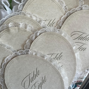 May include: A collection of off-white, oval-shaped table cards with delicate lace edging. Each card features the word "Table" followed by a number in elegant script. The cards are arranged in a slightly overlapping pattern, suggesting a wedding or event setting.