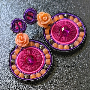 Könnte beinhalten: Ein Paar handgefertigte Ohrringe in einem rosa und violetten Farbschema. Die Ohrringe haben ein kreisförmiges Design mit mehreren Schichten aus Perlen und Stoff. Jeder Ohrring ist mit einer kleinen rosa Blume verziert.
