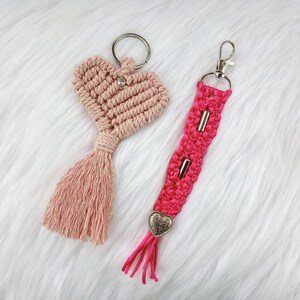 May include: Two handmade macrame keychains. One is a pink heart with a tassel and the other is a pink and silver keychain with a heart charm.