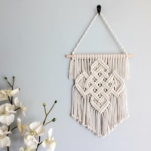 May include: A white macrame wall hanging with a geometric pattern. The hanging is made of cotton cord and is attached to a wooden dowel rod.