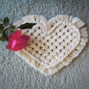 May include: A white crocheted heart-shaped coaster with fringe trim. The coaster is on a light blue fuzzy surface with a pink rose.