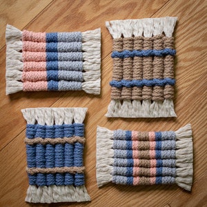 May include: Four handmade macrame coasters with fringe edges. The coasters feature a variety of color combinations, including pink, blue, brown, and gray.