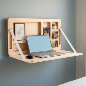 Op de afbeelding: Een aan de muur gemonteerd houten bureau met een lichtgekleurd ontwerp. Het heeft een laptop, muis en opbergvakken voor pennen, een kleine plant en andere items. Witte steunbanden zijn zichtbaar. Het bureau is aan een blauwe muur bevestigd.