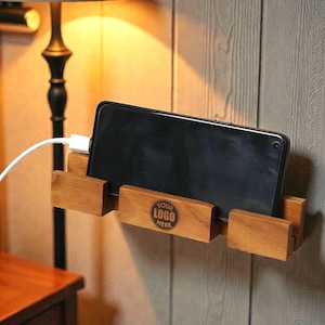 Puede incluir: Soporte de teléfono de madera montado en una pared, con un smartphone negro dentro. El soporte es de madera marrón claro y tiene el texto "YOUR LOGO HERE" impreso. Un cable de carga blanco está conectado al teléfono.