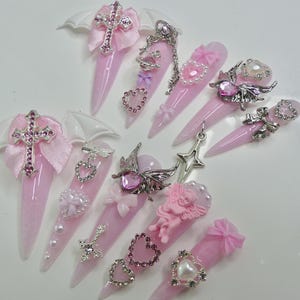 May include: Pink and white nail art featuring a variety of charms, including crosses, hearts, stars, and wings. The charms are silver and have a sparkly finish.
