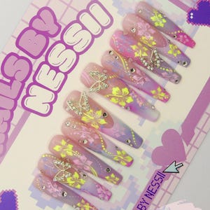 May include: A set of 10 long, pink, purple and yellow ombre press-on nails with a floral and butterfly design. The nails are decorated with rhinestones and gold butterflies. The nails are labelled "Nails By Messi" and "Extra Long".
