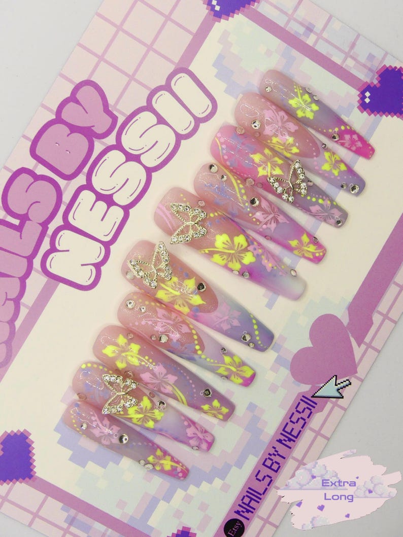 May include: A set of 10 long, pink, purple and yellow ombre press-on nails with a floral and butterfly design. The nails are decorated with rhinestones and gold butterflies. The nails are labelled "Nails By Messi" and "Extra Long".