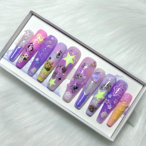 May include: A set of long, coffin-shaped press-on nails in a white box. The nails feature various designs, including cat faces, paw prints, stars, and hearts, in shades of purple, pink, and yellow. Some nails have glitter and pearl accents.