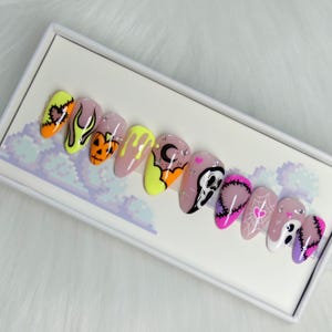 Puede incluir: Un conjunto de uñas postizas en forma de almendra con diseños de Halloween. Las uñas presentan una variedad de colores, incluyendo amarillo neón, naranja, rosa y morado, con diseños como llamas, calabazas, fantasmas y telarañas. Algunas uñas tienen detalles de pedrería.