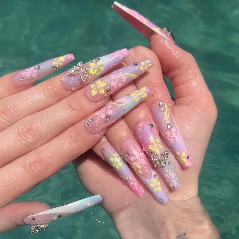 May include: Long, pink and white acrylic nails with a floral design and butterfly accents. The nails are decorated with rhinestones and have a gradient effect.