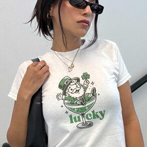 May include: White t-shirt with a green graphic of a leprechaun in a cocktail glass holding a four-leaf clover. The word "lucky" is printed below the glass. The shirt is labeled "White in XS".