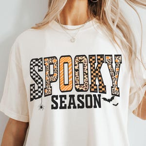 Może przedstawiać: Kremowa koszulka z napisem "Spooky Season" w połączeniu kratki, panterki i pomarańczowych liter. Poniżej tekstu znajduje się pająk i dwa nietoperze. Koszulka ma luźny kr&oacute;j.