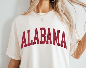 May include: Cream-colored t-shirt with the word "ALABAMA" printed in maroon block letters across the chest. The shirt has short sleeves and a relaxed fit, suitable for casual wear.