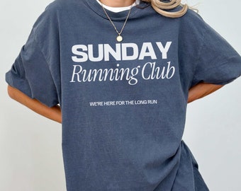May include: A dark blue oversized t-shirt with the text "SUNDAY Running Club" and the words "WE'RE HERE FOR THE LONG RUN" in white. The t-shirt is made of a soft, comfortable material.