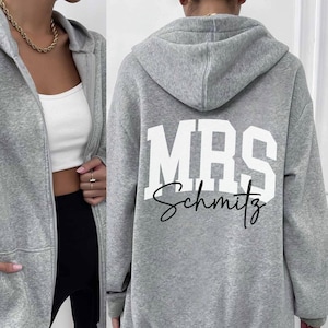 May include: A light gray zip-up hoodie with a hood and front pockets. The back of the hoodie features the text "MRS Schmitz" in white block letters and script. The hoodie is made of a soft, comfortable material.