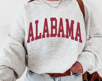 May include: Ash grey sweatshirt with the word "ALABAMA" in maroon block letters across the chest. The sweatshirt has long sleeves and a crew neck. The text "Ash Grey" is at the bottom left corner.