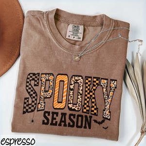 Może przedstawiać: Brązowa koszulka z napisem "SPOOKY SEASON" w mieszance wzor&oacute;w i kolor&oacute;w. Koszulka ma małą grafikę pająka i sylwetki nietoperzy. Na zdjęciu znajduje się r&oacute;wnież srebrny naszyjnik z wisiorkiem i brązowy kapelusz.
