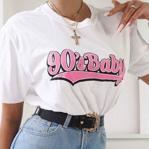 May include: A white t-shirt with a pink graphic that says "90's Baby" in a retro font. The shirt is paired with light blue jeans and a black belt with a gold buckle.