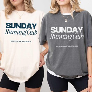 Puede incluir: Dos camisetas con el texto "SUNDAY Running Club" en diferentes colores. La camiseta de la izquierda es de color marfil con texto azul marino. La camiseta de la derecha es gris con texto blanco. Ambas camisetas tienen el texto "WE'RE HERE FOR THE LONG RUN" debajo del texto principal.