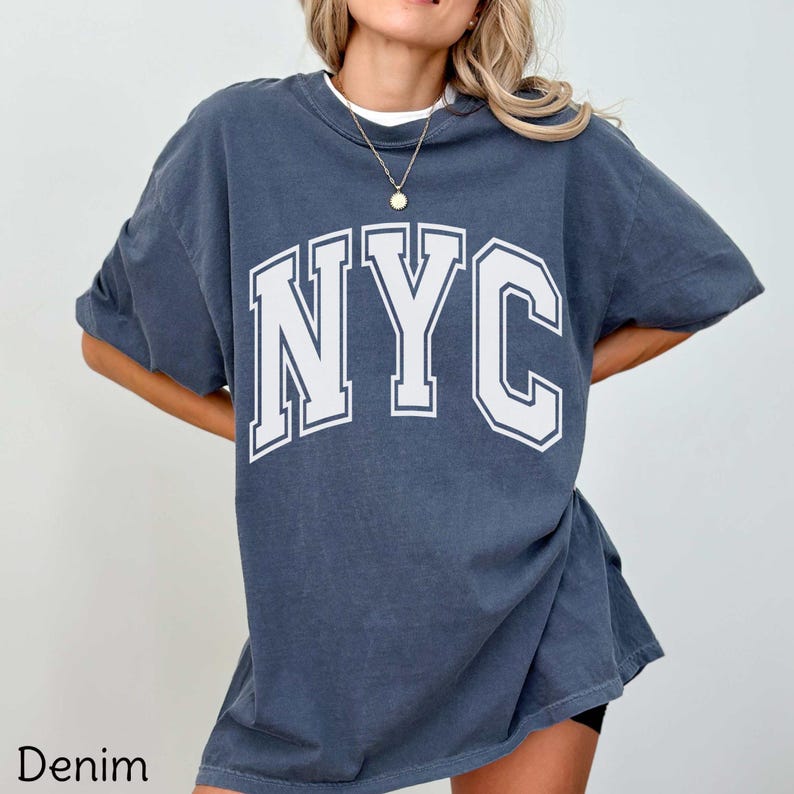 May include: A denim-colored oversized t-shirt with the white letters "NYC" in a collegiate font. The shirt has short sleeves and a crew neck. The word "Denim" is printed at the bottom left corner.