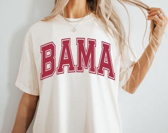 May include: Cream-colored t-shirt with the word "BAMA" in maroon block letters with a white outline. The shirt has short sleeves and a relaxed fit, suitable for casual wear.