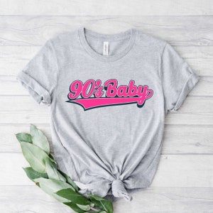 May include: Grey t-shirt with a pink and blue graphic that says "90's Baby" in a retro font.