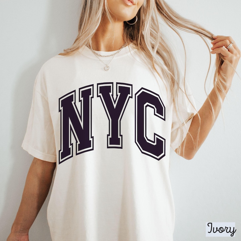 May include: Ivory-colored t-shirt with the letters "NYC" in a dark purple collegiate font. The shirt has a crew neck and short sleeves. The word "Ivory" is printed in a small, white font in the bottom right corner.
