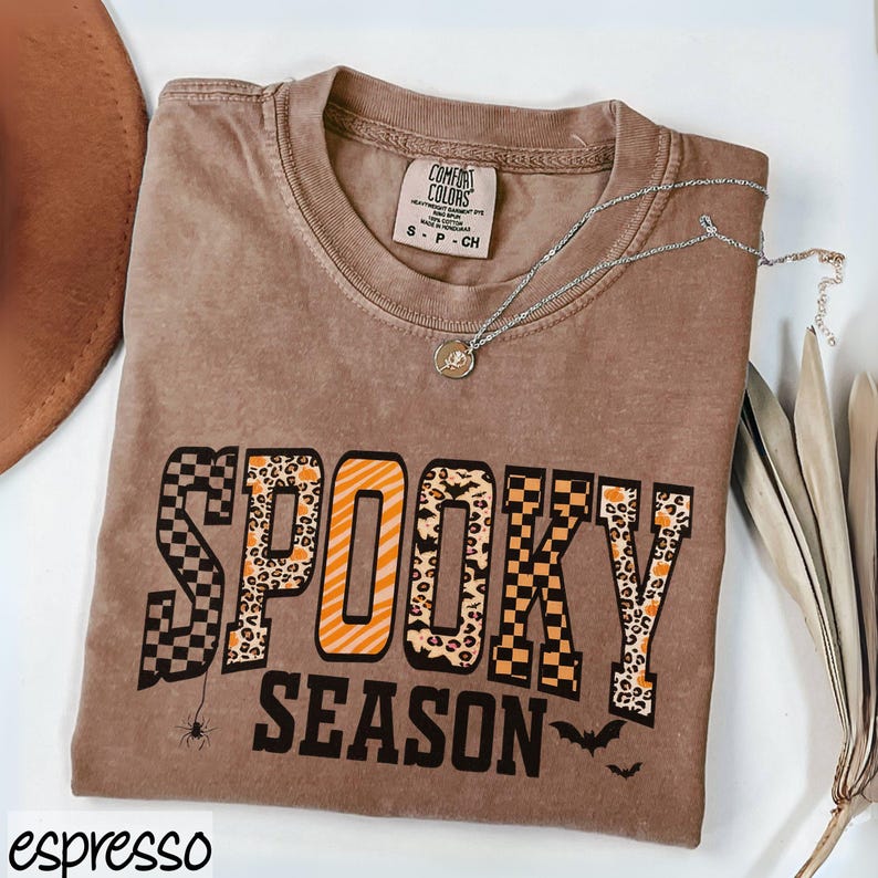 Może przedstawiać: Brązowa koszulka z napisem "SPOOKY SEASON" w mieszance wzor&oacute;w i kolor&oacute;w. Koszulka ma małą grafikę pająka i sylwetki nietoperzy. Na zdjęciu znajduje się r&oacute;wnież srebrny naszyjnik z wisiorkiem i brązowy kapelusz.