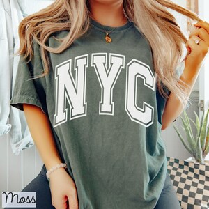 May include: Dark green t-shirt with the white letters "NYC" in a collegiate font. The shirt is a relaxed fit and has short sleeves. The text is outlined in a contrasting color.