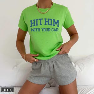 Puede incluir: Una camiseta verde lima con el texto "HIT HIM WITH YOUR CAR" en letras azules. La modelo lleva la camiseta con pantalones cortos grises.
