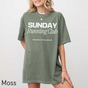 Puede incluir: Una camiseta de gran tama&ntilde;o de color verde musgo con el texto "SUNDAY Running Club" y las palabras "WE'RE HERE FOR THE LONG RUN" en blanco.
