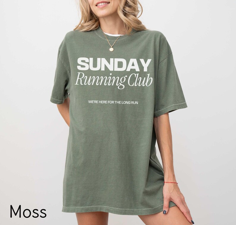 Puede incluir: Una camiseta de gran tama&ntilde;o de color verde musgo con el texto "SUNDAY Running Club" y las palabras "WE'RE HERE FOR THE LONG RUN" en blanco.