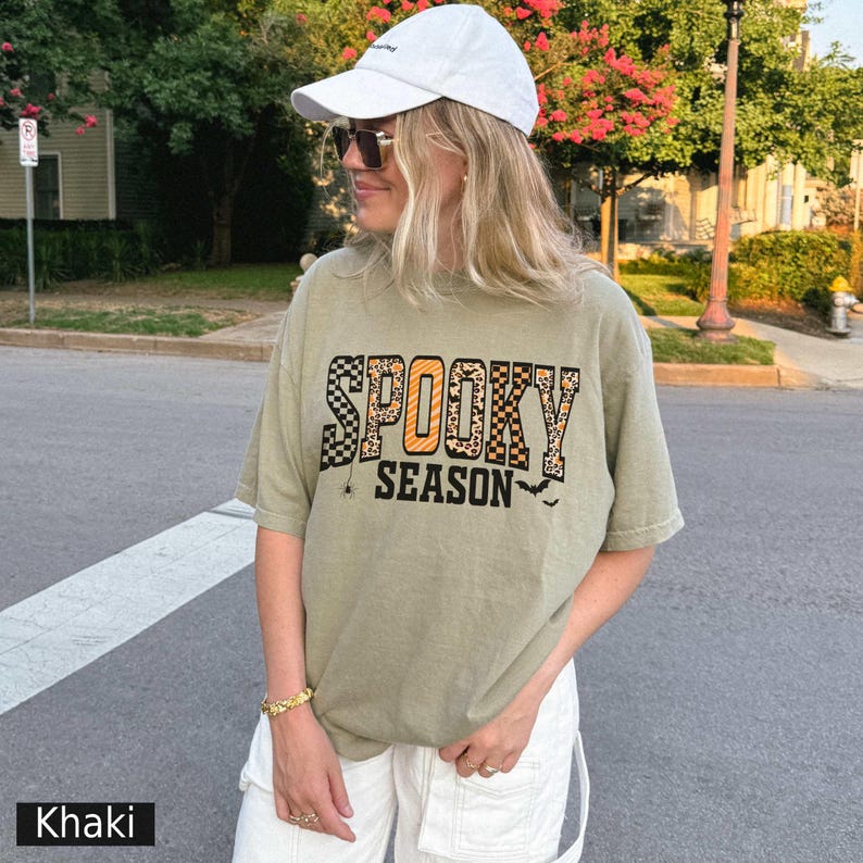 Może przedstawiać: Koszulka w kolorze khaki z napisem "SPOOKY SEASON" w połączeniu z kratą, panterką i pomarańczowymi literami. Koszulka posiada r&oacute;wnież małą grafikę pająka i nietoperza. Osoba nosi białą czapkę z daszkiem i okulary przeciwsłoneczne.