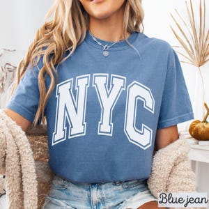 May include: Blue jean colored t-shirt with the white text "NYC". The shirt has short sleeves and a crew neck. The text is in a bold, block letter style. The shirt is made of a soft material.