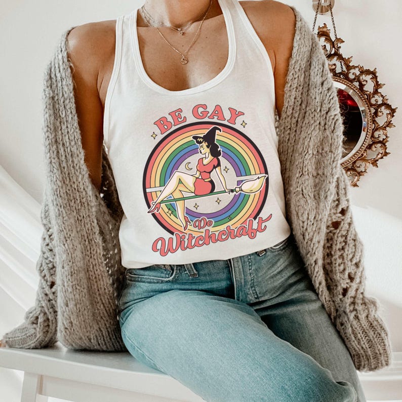 May include: White tank top with a rainbow and a witch flying on a broomstick. The text on the shirt says "Be Gay Do Witchcraft".